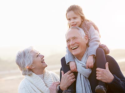 An old man holding his granddaughter on his shoulders with his wife standing next to him smiling